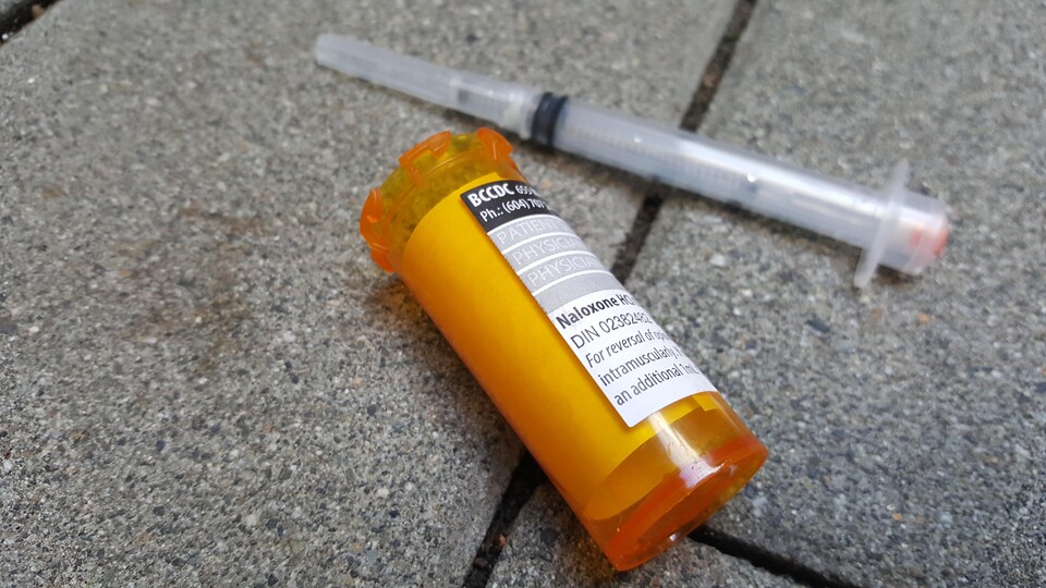 Une seringue usée et un flacon ouvert sur lequel est apposé une ordonnance pour naloxone sont sur le pavé.