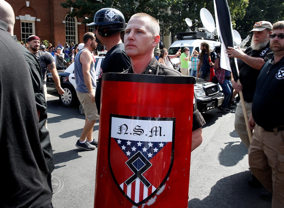 Un homme porte un bouclier rouge avec l'insigne du Nationalist Socialist Movement