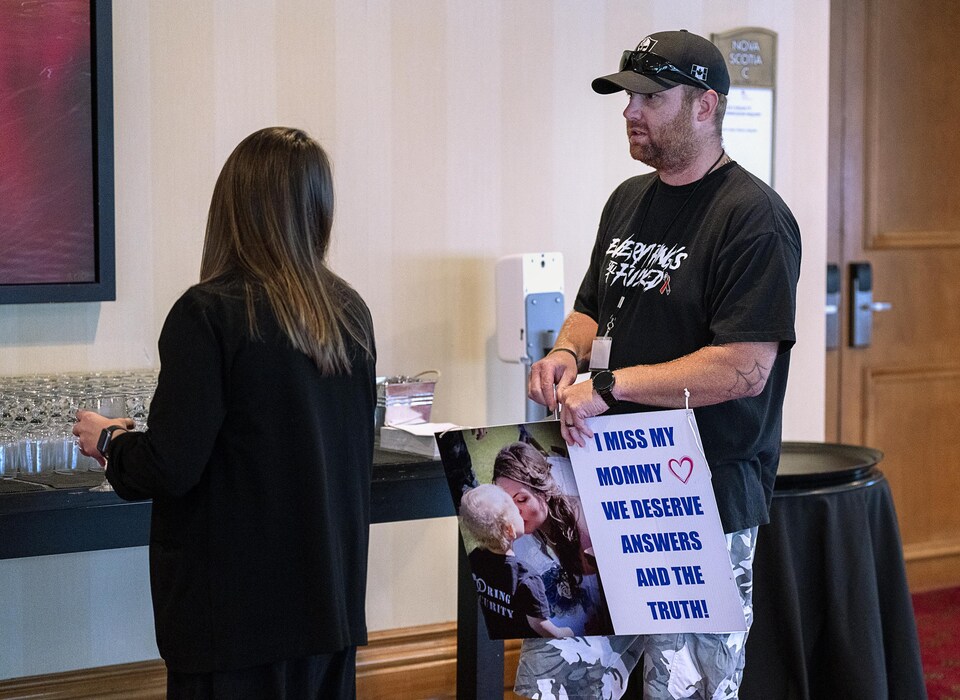 Nick Beaton au centre des congrès. Il parle à une femme de dos. Il tient une affiche sur laquelle on voit une photo de sa conjointe Kristen Beaton et de leur enfant, ainsi que la phrase «Je m'ennuie de ma maman. Nous méritons des réponses et la vérité.»
