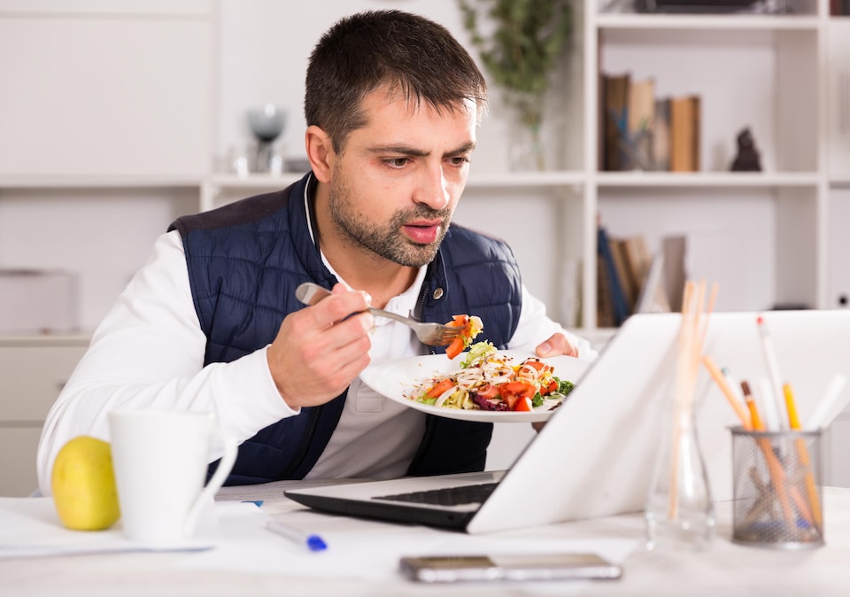 Un homme tient son assiette et mange devant son ordinateur portable.