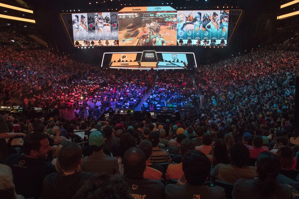 Une foule de plusieurs milliers de personnes massés dans les gradins d'un aréna regardent une scène et des écrans géants affichant une partie du jeu vidéo Overwatch.