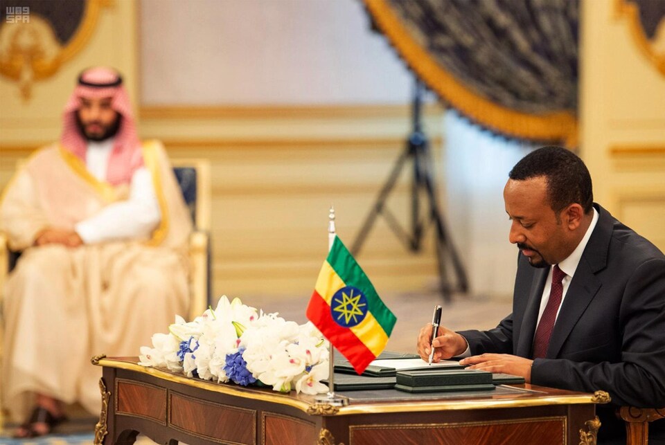Un homme assis, signe un papier. Derrière lui ont aperçoit un haut dignitaire saoudien.