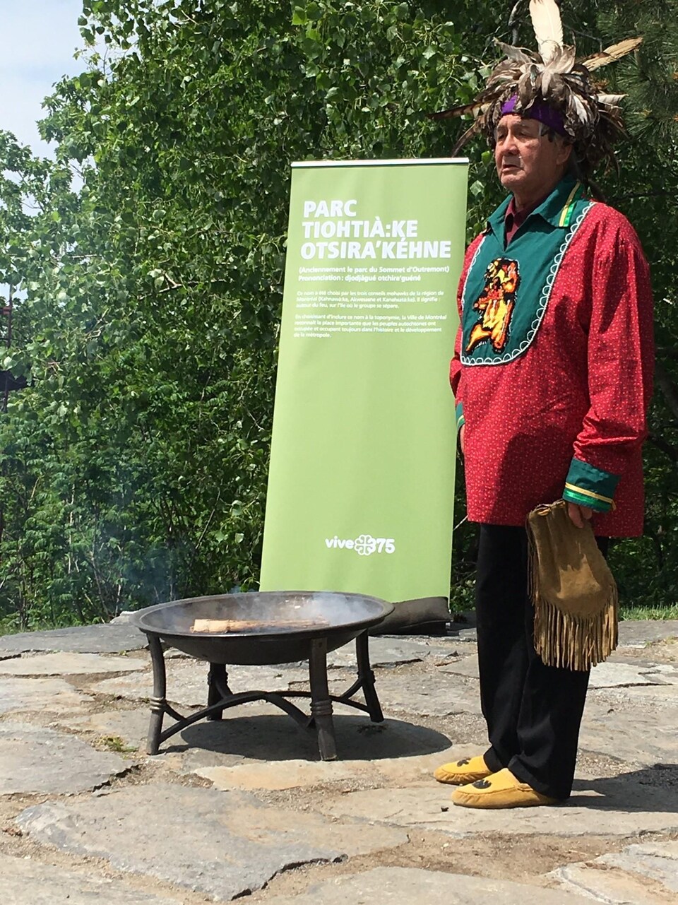 Un Autochtone vêtu d'habits traditionnels, comme une petite coiffe de plume, se tient devant une affiche montrant le nouveau nom du parc au sommet d'Outremont.