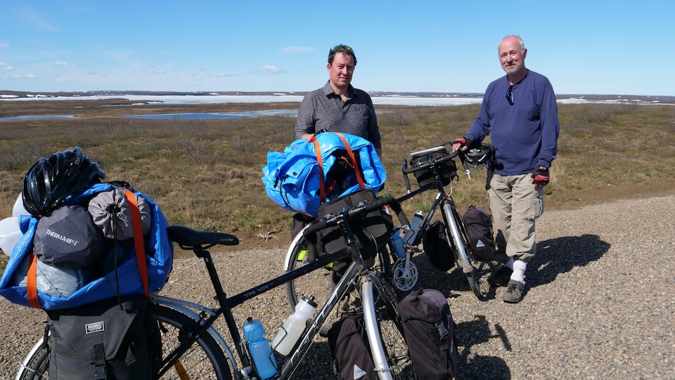 Deux hommes et leurs vélos sur une route de gravier devant la toundra où se trouvent de petits lacs et des résidus de neige.