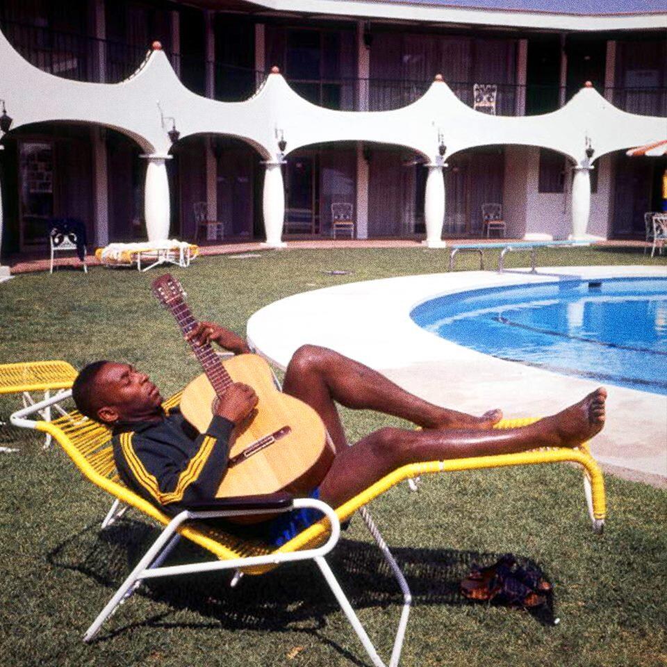 Un homme est étendu sur une chaise longue au bord d'une piscine extérieure, guitare dans les mains. 