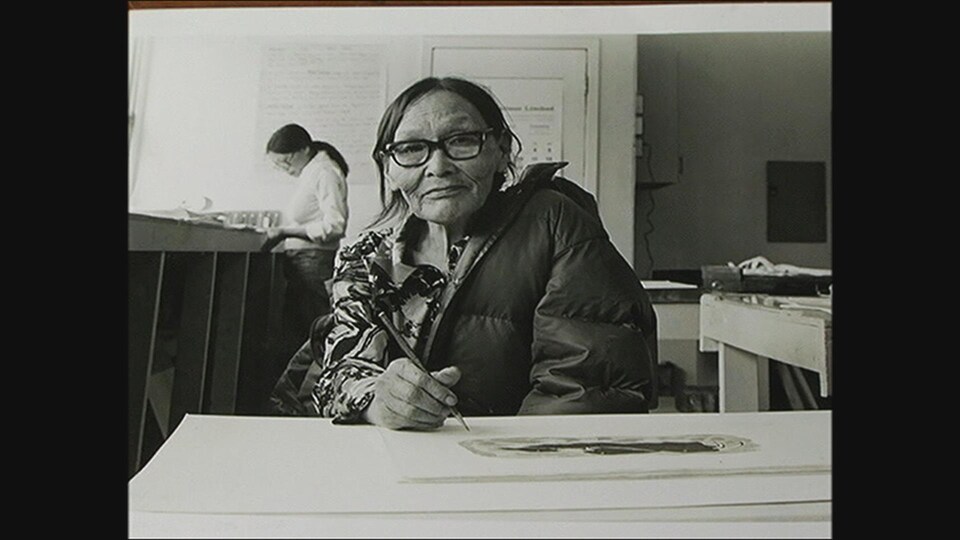 Une femme inuite, vêtue d'un manteau, semble prendre une pause de son dessin le temps que le photographe prenne la photo. Elle porte des lunettes à forte monture et tient un crayon en main. 