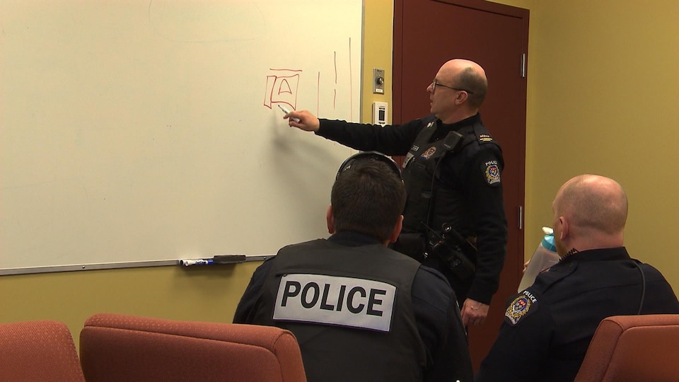 Un policier fait un plan au tableau sous l'oeil attentif de ses deux collègues.