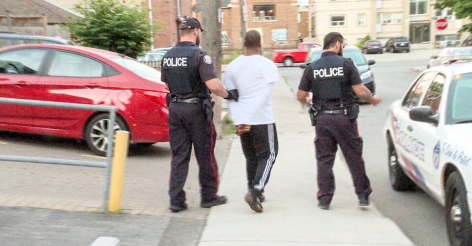 Photo d'un homme, les mains menottées derrière le dos, qui est amené par deux policiers vers une autopatrouille.
