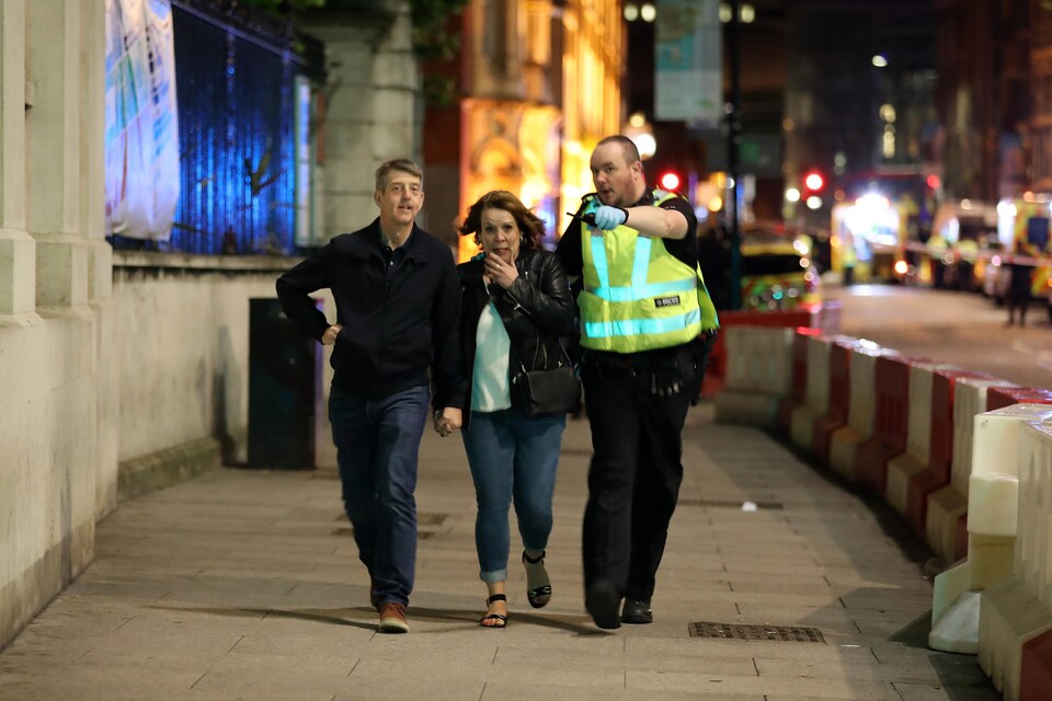 Un policier portant un dossard jaune et des gants de plastique bleus pointent devant lui, semblant diriger un homme et une femme vers un lieu sûr.