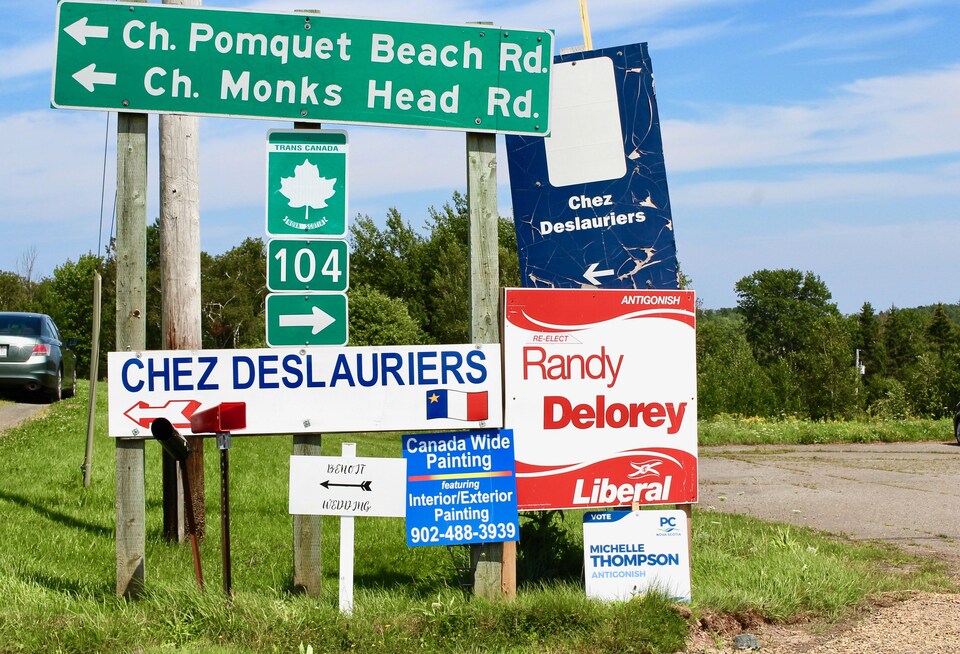 Un panneau routier indiquant la direction pour la plage de Pomquet avec autour des affiches électorales.