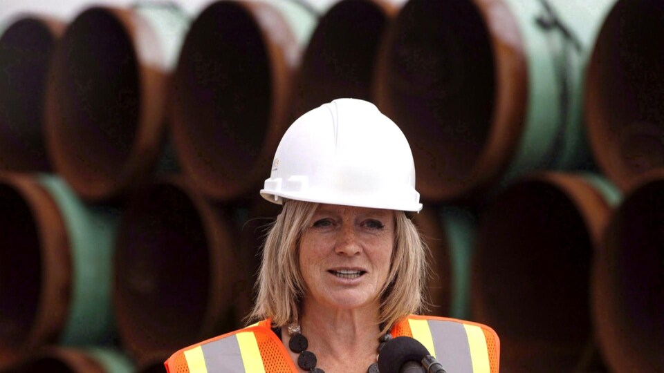 Rachel Notley qui porte un casque de construction devant des tuyaux de pipeline. 