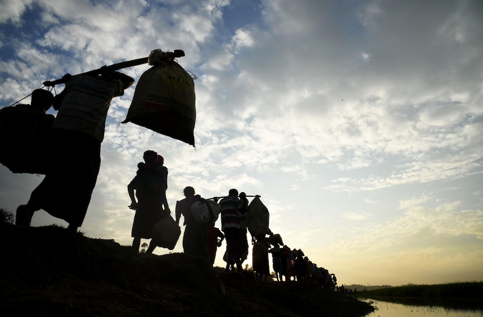 Des familles de rohingyas transportent leurs biens et leurs enfants. Elles marchent en file, en longeant un cours d'eau.