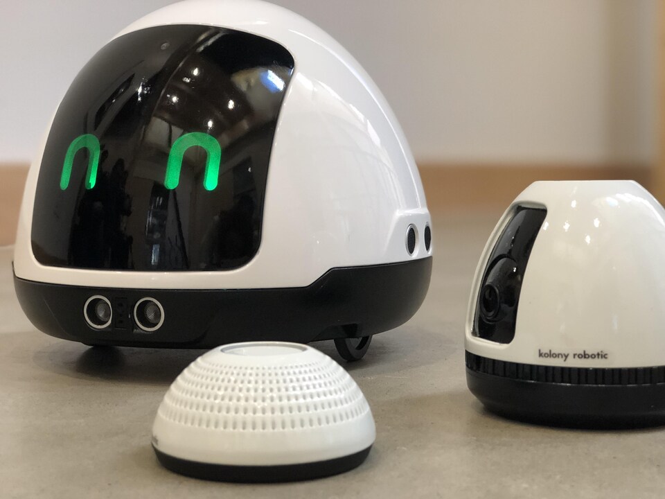 Un robot noir et blanc est accompagné d'une caméra et d'un haut-parleur. Le robot à des yeux verts et il est sur roulette.