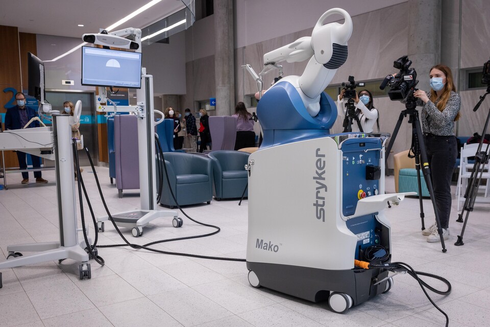 Une machine rectangulaire blanche et bleue sur roues est coiffée d'un bras robotique et reliée par des câbles à un écran. Des gens sont attroupés autour de l'appareil avec des caméras sur trépied pour en prendre des images.