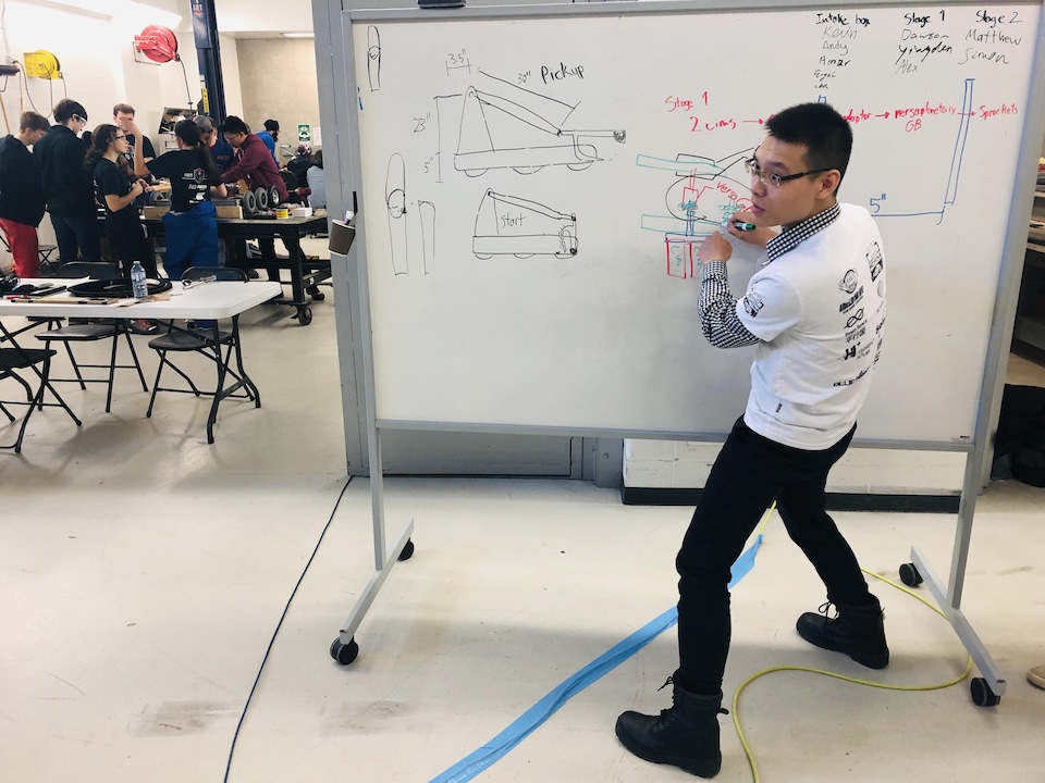 Dans le fond, à gauche, des étudiants montent le robot, au premier plan, à droite, un étudiant dessine sur un tableau blanc le plan du bras mécanique du robot.