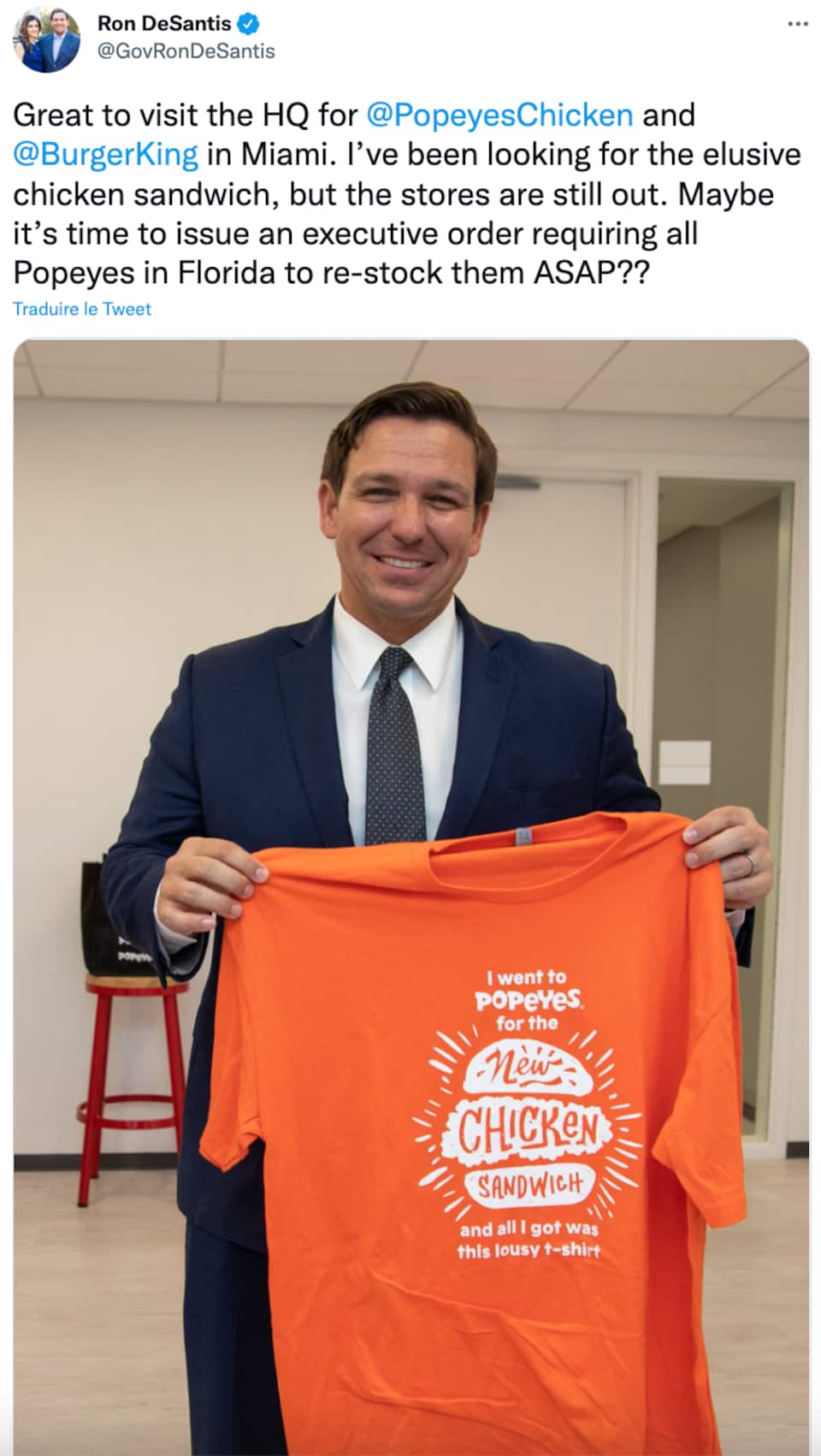 On voit un tweet de Ron DeSantis; c'est une photo où il sourit à la caméra et tient dans ses mains un t-shirt orange avec un logo en lettres blanches faisant la promotion d'un restaurant de poulet frit.