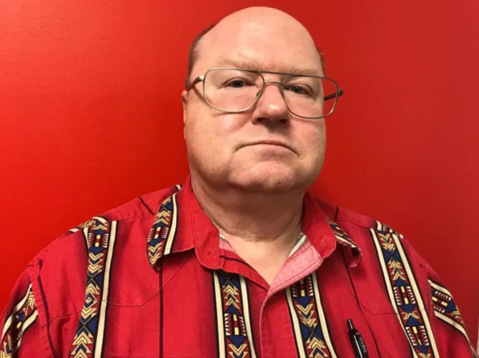 Le maire de La Ronge porte des lunettes, une chemise rouge à motifs. Derrière lui, le décor est rouge vif. 
