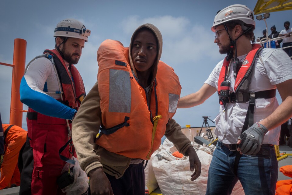 Un jeune homme africain portant une veste de sauvetage est flanqué de deux hommes travaillant pour SOS Méditerranée sur le pont de l'Aquarius.