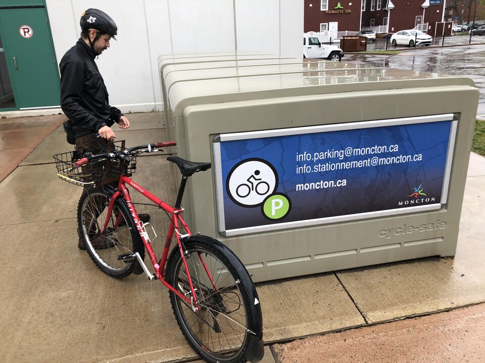 Un cycliste debout avec son vélo, à côté d'un casier à vélo qui sert à stationner son vélo.