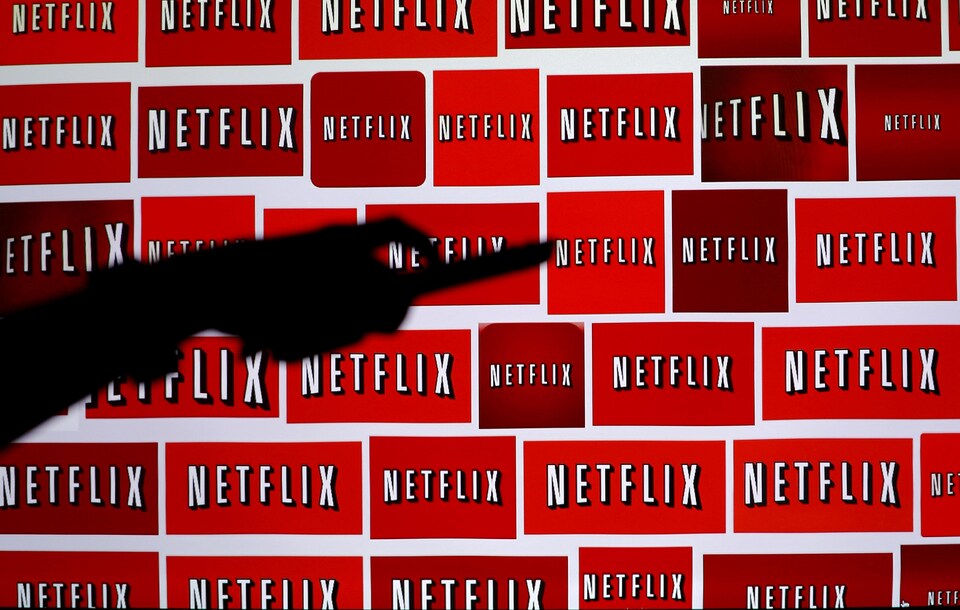 Une main tenant une télécommande devant une série de logos du service Netflix.