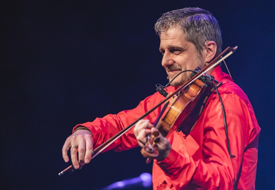 Un musicien sur scène photographié de profil, vêtu d'une chemise rouge et jouant du violon.