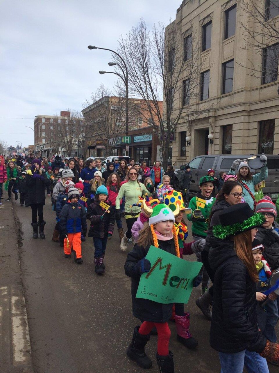 Des dizaines d'adultes et d'enfants dont certains tiennent de petits drapeaux fransaskois marchent dans une rue.