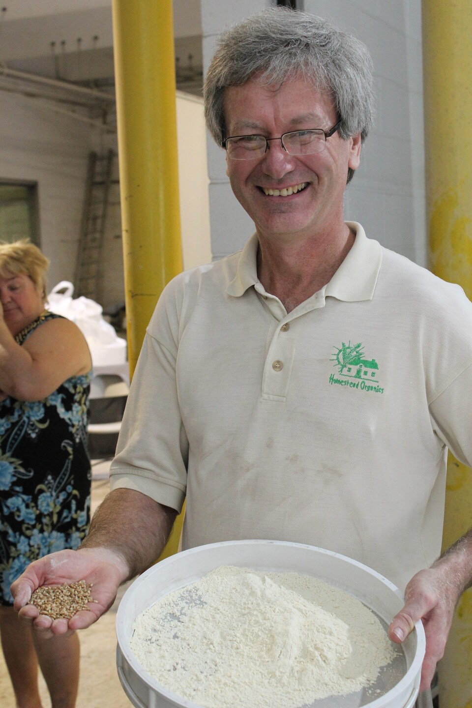 Tom Manley sourit à la caméra; il tient un seau de farine dans une main et des graines dans l'autre.