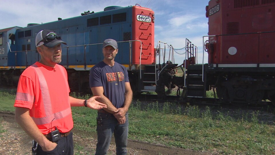 Deux hommes discutent devant un train, à l'extérieur.