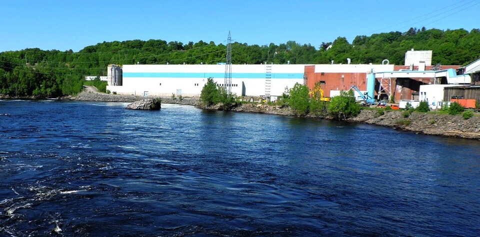 On voit l'usine (un grand bâtiment blanc de forme rectangulaire) au bord de la rivière Saint-Maurice.