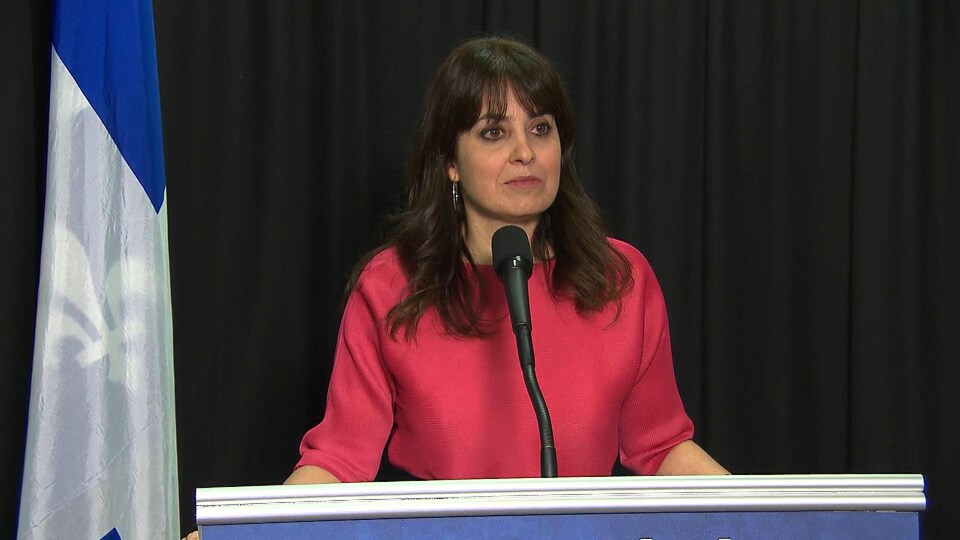 Une dame aux cheveux bruns et aux yeux bruns est vêtue d'un chandail rose et se tient sur un podium.