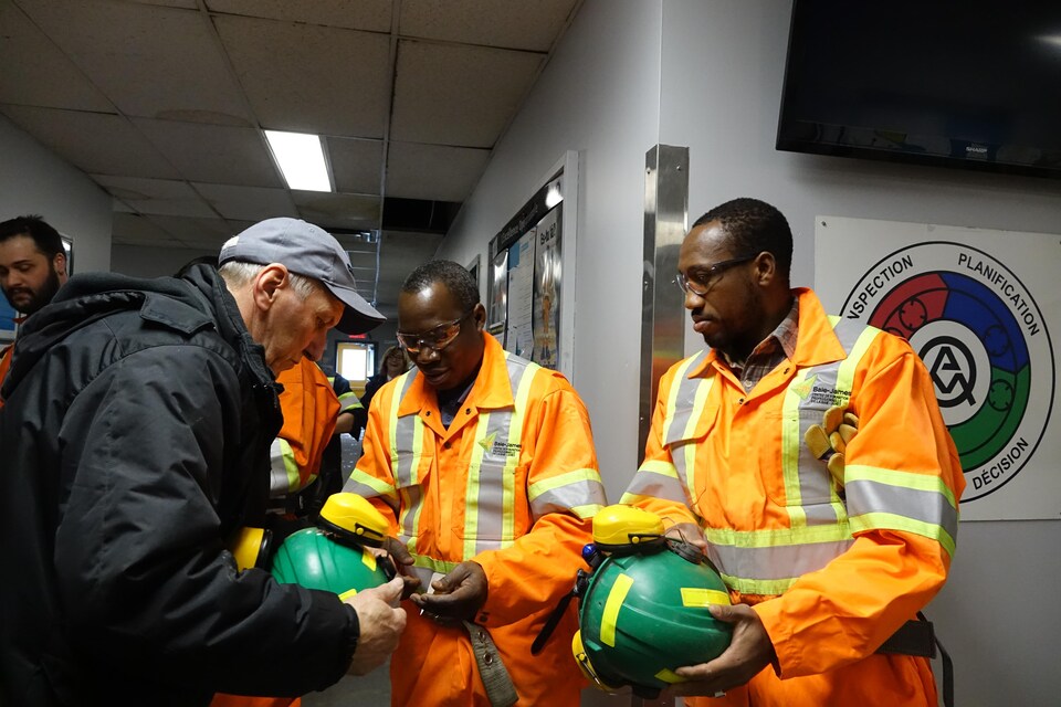 Deux candidats au DEP habillés en combinaison de mineur orange se font expliquer comment mettre leur casque vert, avant de descendre dans la mine.