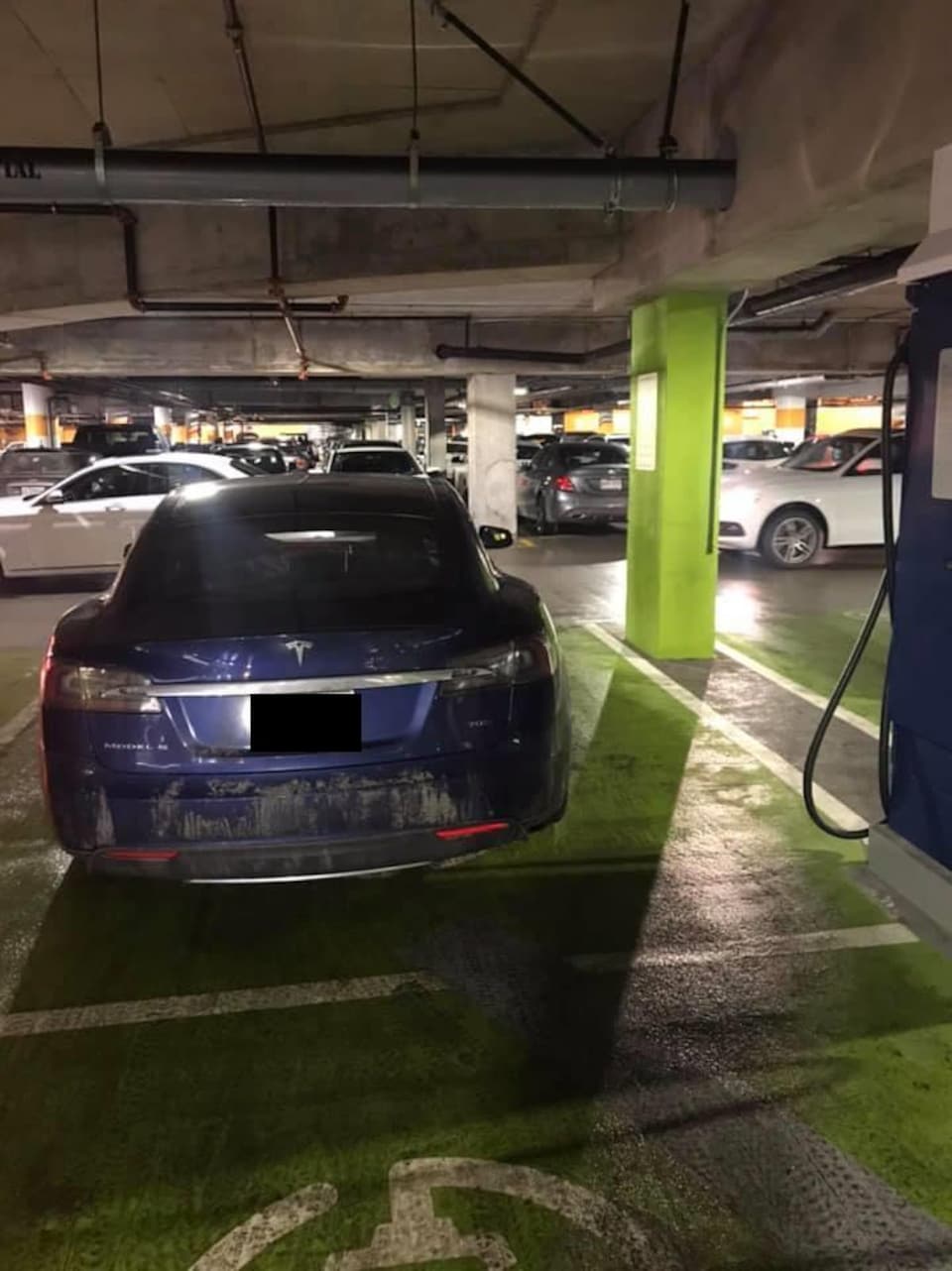 Un véhicule électrique bleu de marque Tesla occupe un espace de stationnement réservé peint en vert, mais il n'est pas branché à la borne de recharge à sa droite.