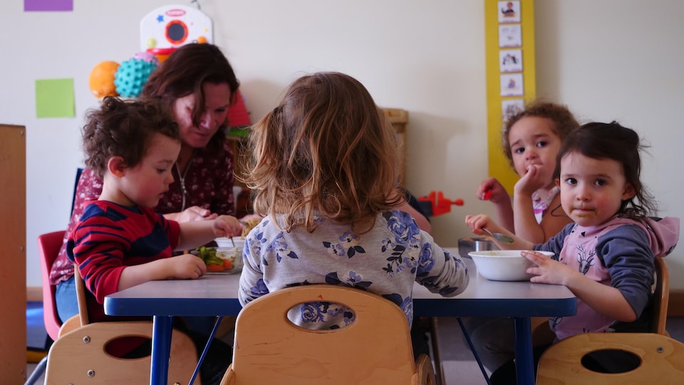 Des enfants et une éducatrice assis sur de petites chaises en bois à une table mangent.