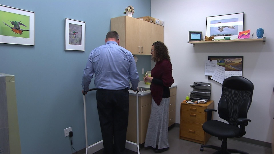 Un homme est debout sur une balance à côté d'une femme qui prend des notes dans un cabinet médical.