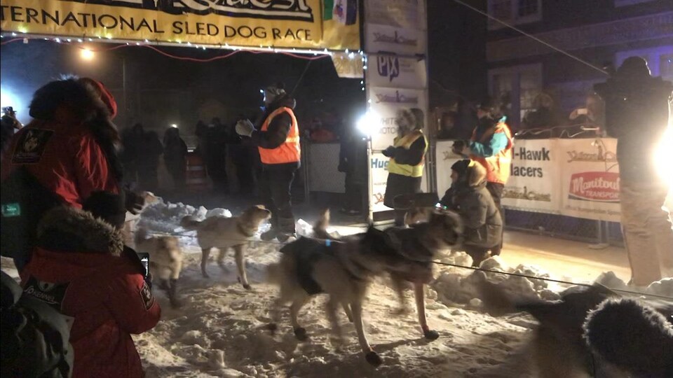 Des bénévoles applaudissent et des journalistes ainsi que le public prennent des photos ou des vidéos alors qu'un premier meneur de chiens franchit la ligne d'arrivée le soir sur la neige. Tous sont habillés très chaudement.