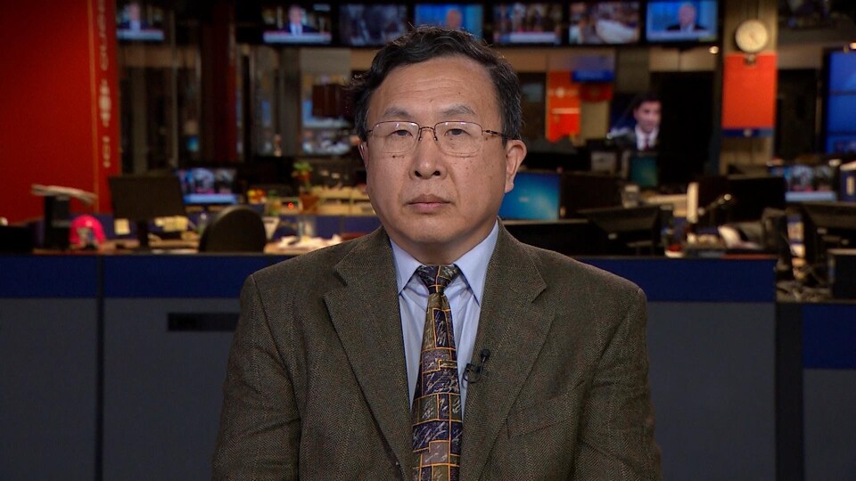 Un homme asiatique portant des lunettes, un veston et une cravate, regarde la caméra sans sourire.