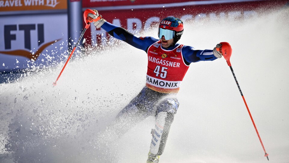 Le skieur AJ Ginnis, entouré de neige, lève ses bras à l'arrivée d'un parcours.