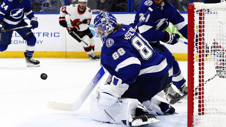 Le gardien du Lightning, en bleu, à genou devant son filet, surveille la rondelle en plein vol devant lui. 