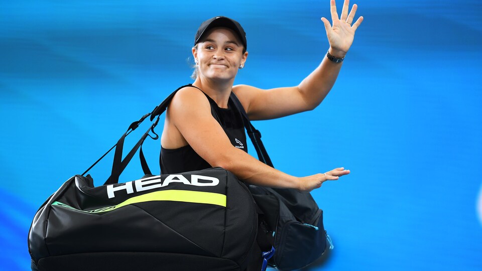 Une joueuse de tennis fait un signe de la main en portant des sacs