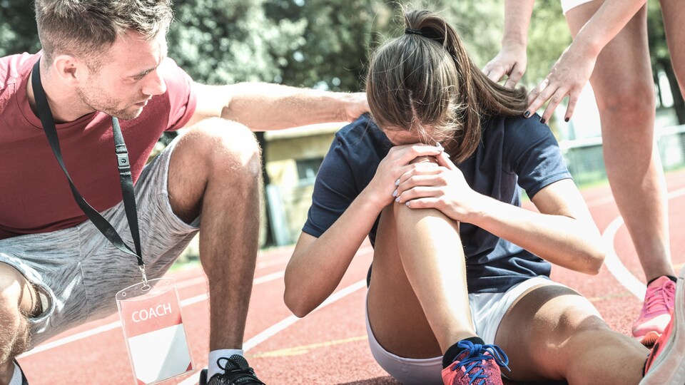 L'adolescente, assise au sol, se tient le genou. Son entraîneur la réconforte.