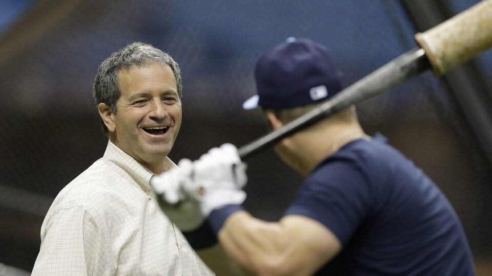 Un homme en chemise blanche sourit en regardant un joueur de baseball prendre un élan à l'entraînement. 