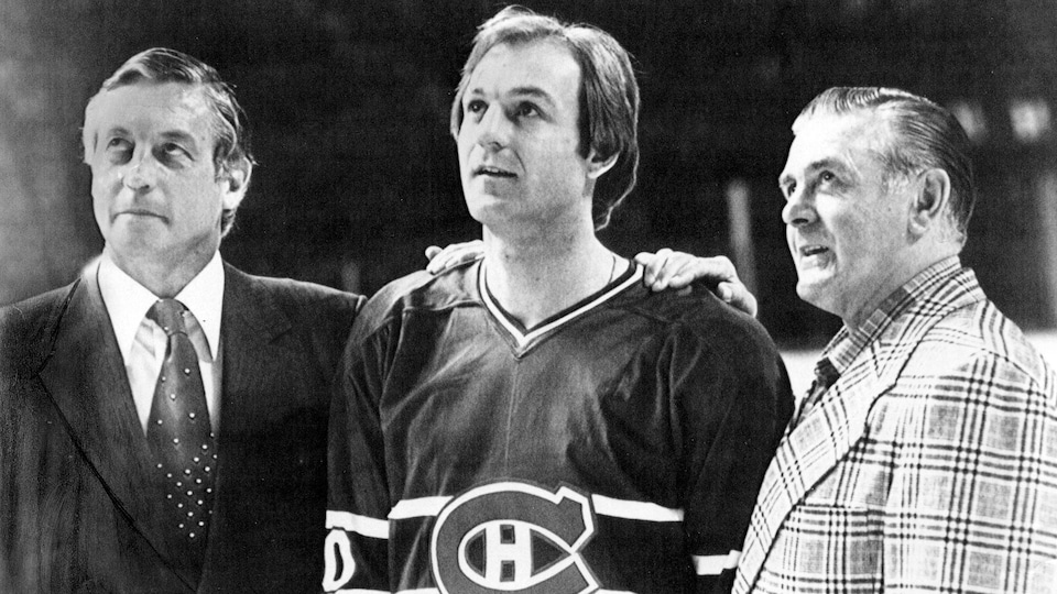 Photo en noir et blanc d'un joueur du Canadien en uniforme, entouré de deux hommes en complet. 