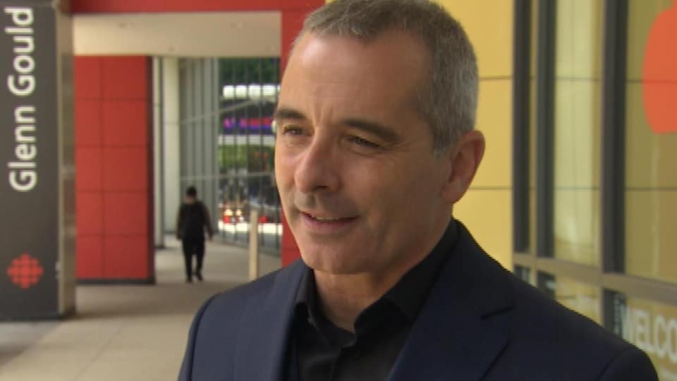 A man gives a TV interview in a hallway with the Radio-Canada logo in the background.