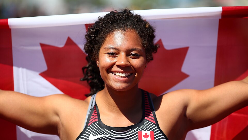 L'athlète de Richmond sourit, le drapeau canadien derrière elle, après avoir gagné l'or aux Championnats U-20 d'athlétisme, en 2018.