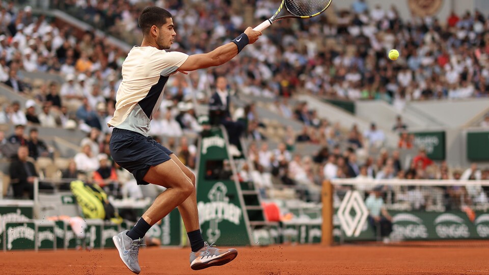 Un joueur de tennis, de dos, renvoie la balle d'un coup droit sur un terrain en terre battue devant des gradins pleins.