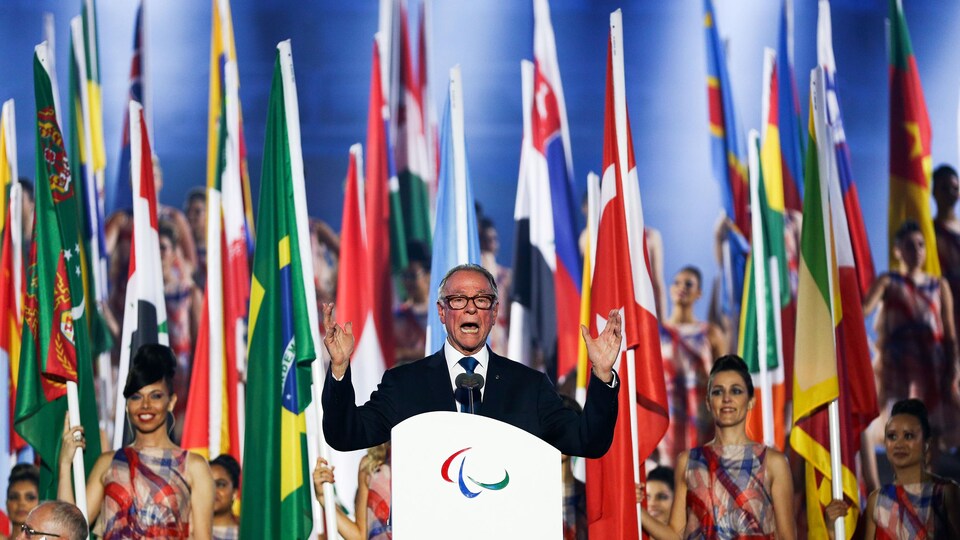 Un homme en veston prononce un discours, debout derrière un podium, les bras au ciel, alors que défilent derrière lui les drapeaux des délégations.