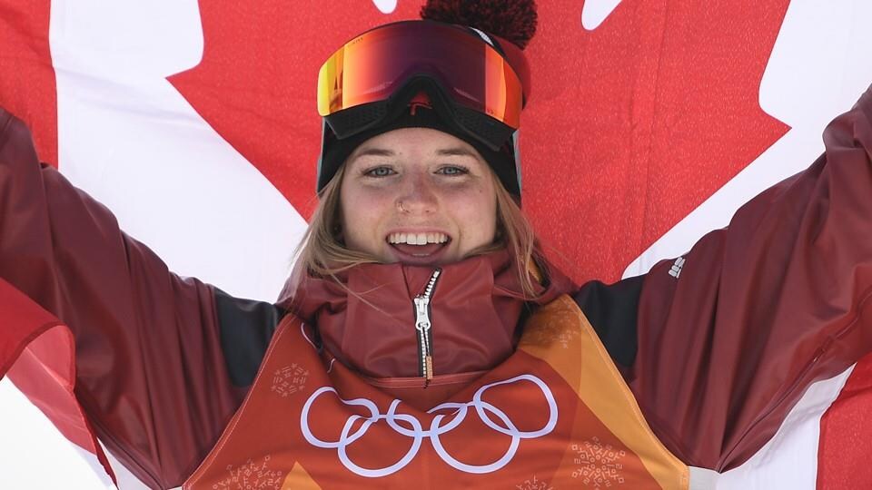 Une athlète souriante avec un dossard olympique tient derrière elle le drapeau canadien.