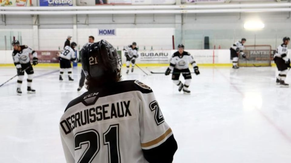 Hockey player, back to camera, with a vest marked 21 and Desruisseaux, looks at the rink and other players who are skating.