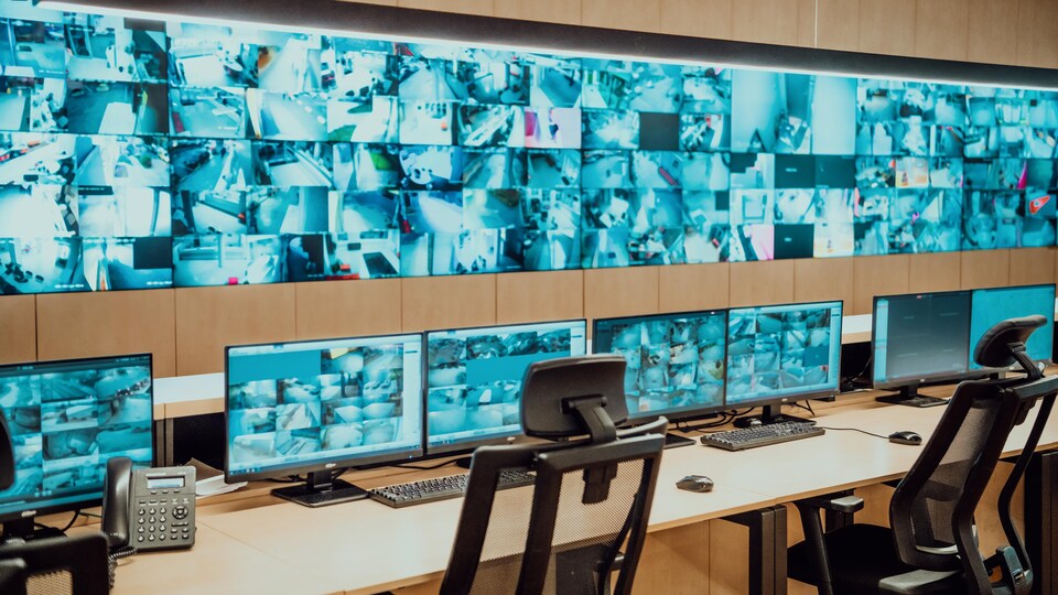 Un énorme écran montre des dizaines d'images captées par des caméras de sécurité. Des ordinateurs et des chaises se trouvent devant.
