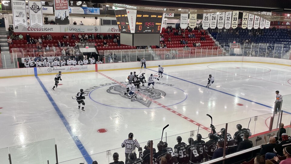 Des joueurs de hockey s'affrontent sur la glace. On voit des gradins avec quelques spectateurs et de nombreuses bannière accrochées au plafond.